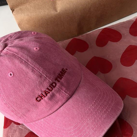 Une casquette en denim rose brodée 'Chaud Time' posé sur un décor fait de coeurs